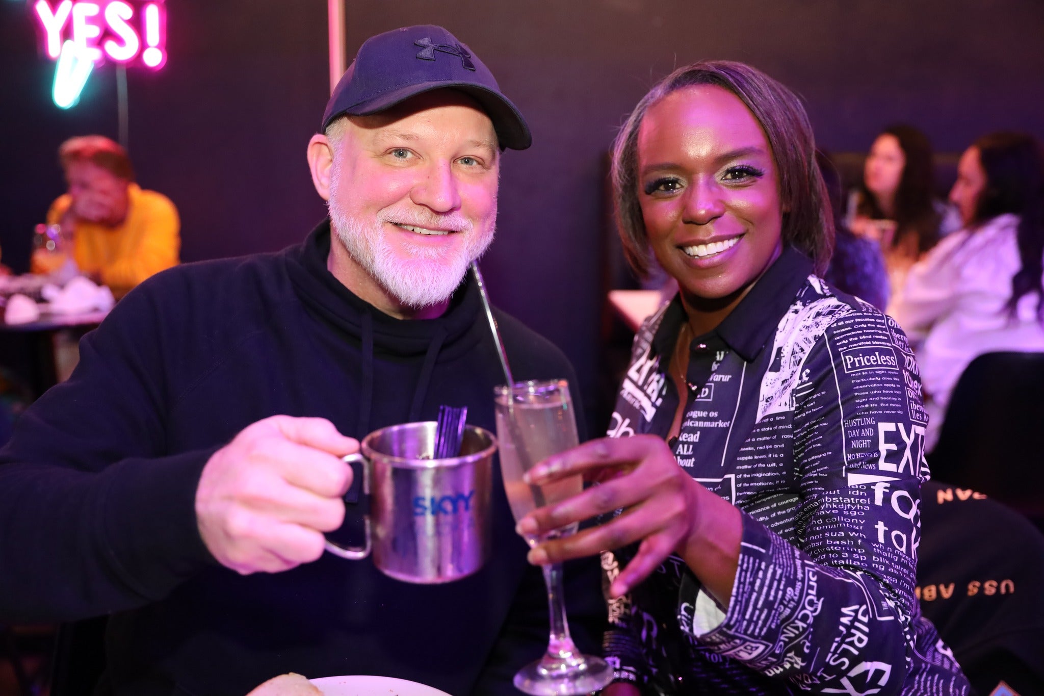 Two people enjoying drinks together at a social event with a neon sign in the background.
