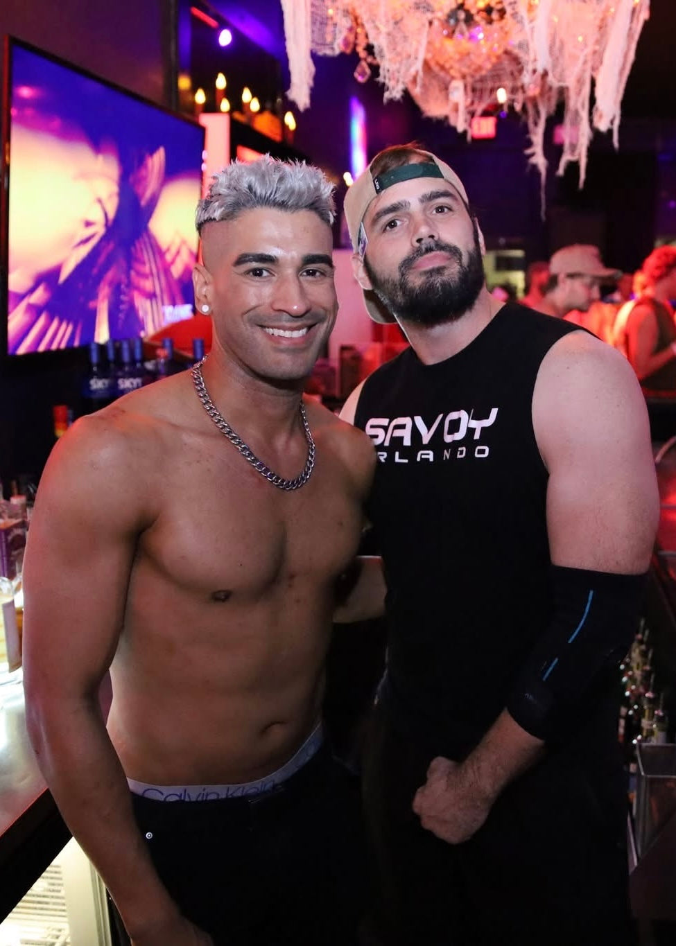 Two men posing together in a nightclub setting with a chandelier and colorful lights.