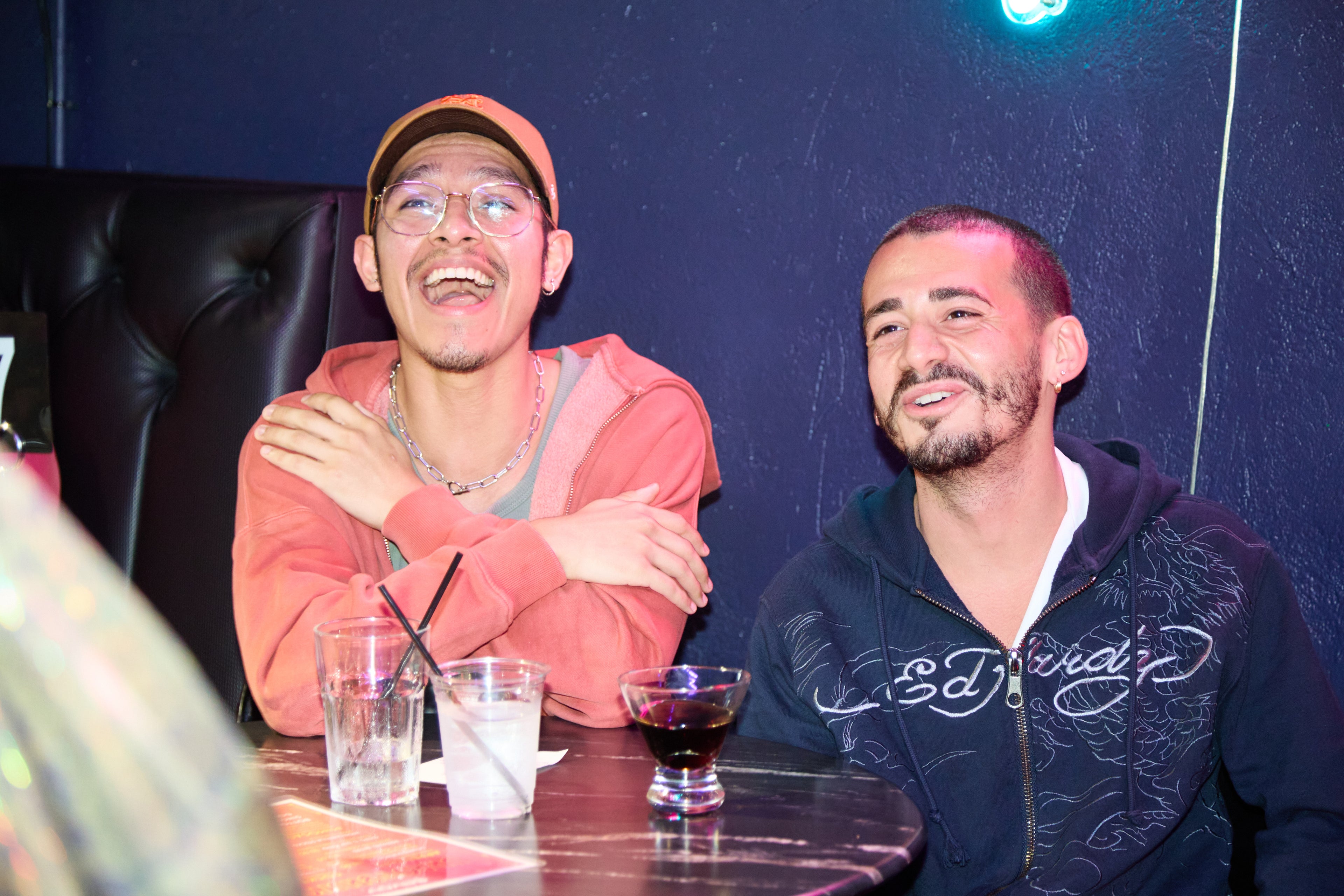 Two men sitting at a table with drinks, one wearing a red hoodie and cap, the other in a dark jacket with 'ED' branding.