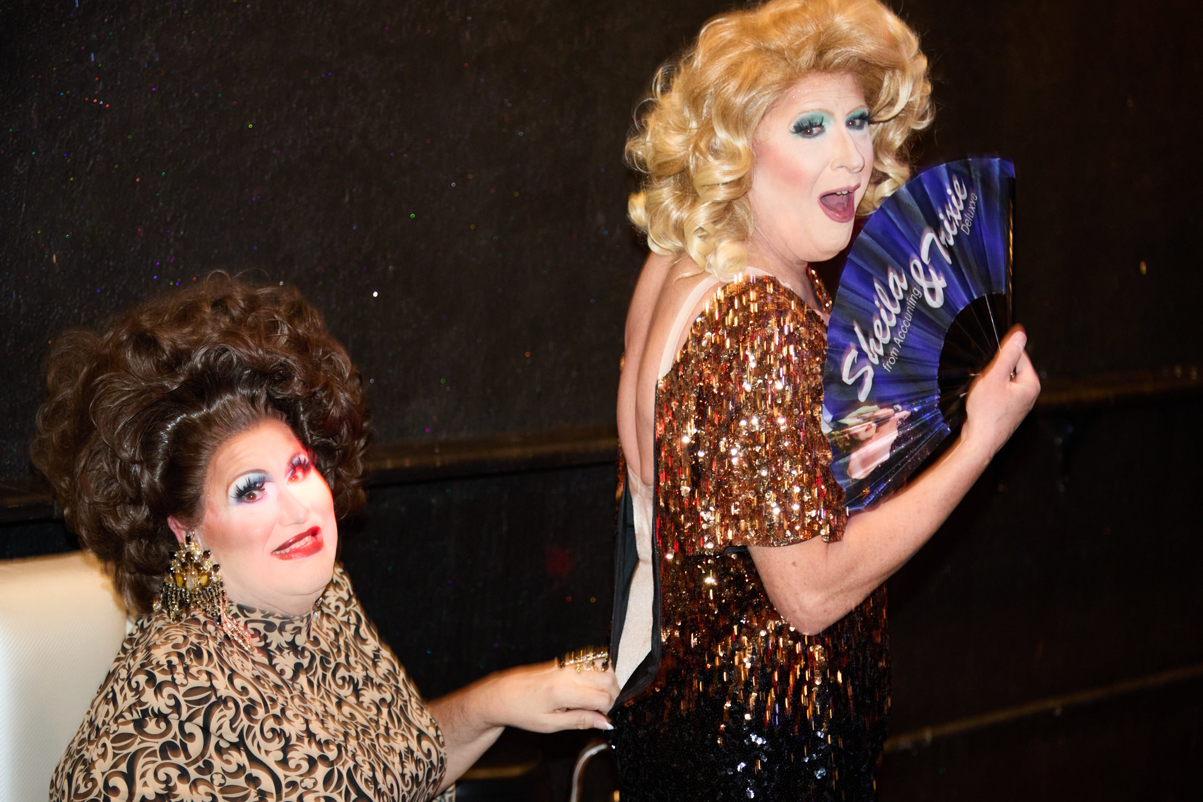 Two drag queens in elaborate costumes with one holding a fan.