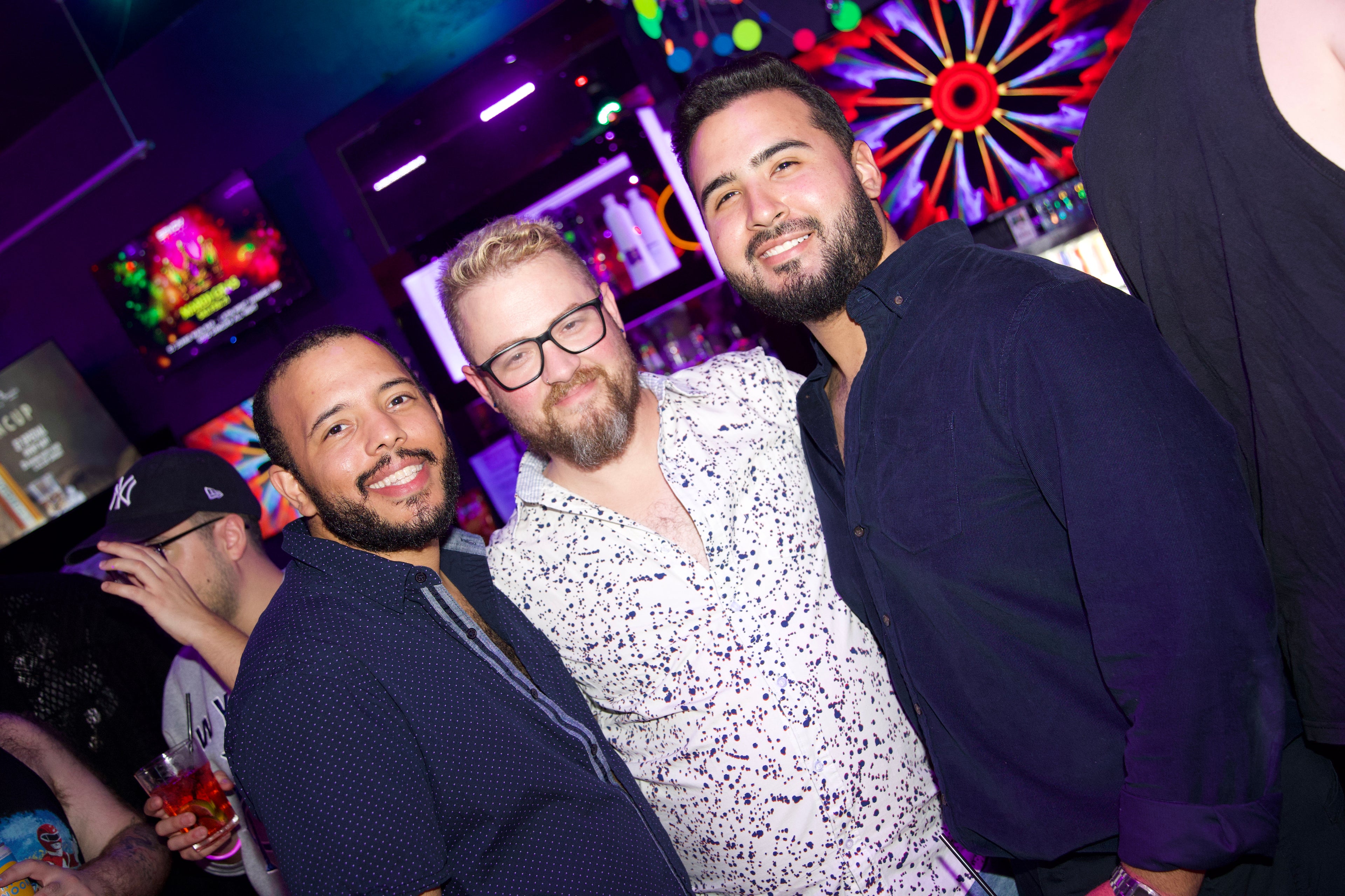 Three men posing together in a lively nightclub setting with colorful lights and decorations.