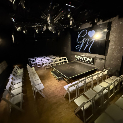 The interior of a theater with rows of chairs and a stage, featuring the Savoy Theatre logo.