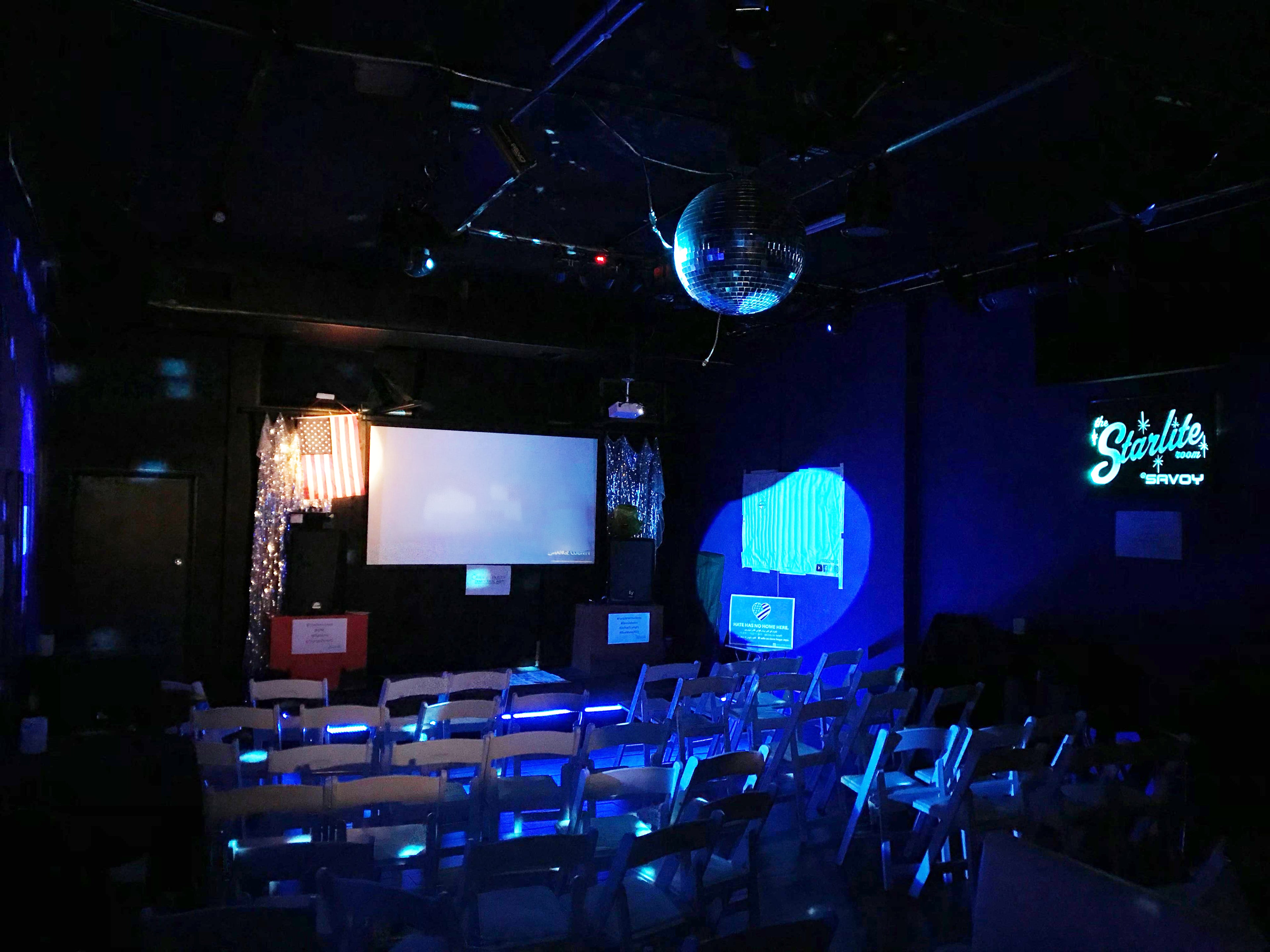Dark room with chairs arranged for an event, screen, disco ball, and neon sign.